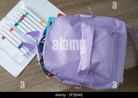 Lila Schulrucksack auf einem Schreibtisch mit Notizbüchern und bunten Stiften, bereit für einen neuen Schultag. Stockfoto