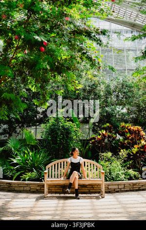 Ansicht einer jungen Frau, die auf einer Holzbank sitzt, umgeben von üppiger Vegetation und tropischen Pflanzen in den Gewächshäusern des Chicago Botanic Garden, Glen Stockfoto