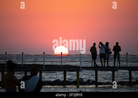 Eine Gruppe von vier jungen Männern ist als Silhouette auf einem hölzernen Küstenpier mit Seilgeländern zu sehen, während sie den Sonnenuntergang am Horizont beobachten. Stockfoto