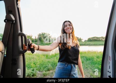 Ansicht einer lächelnden jungen Frau, die neben einer offenen Autotür auf einem grasbewachsenen Feld nahe einem Fluss in Brandon, Manitoba, Kanada, steht. Stockfoto