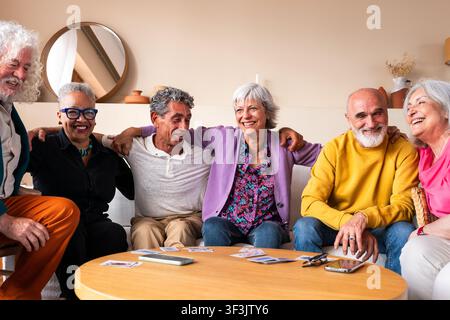 Fröhliche Gruppe älterer Freunde, die sich umarmen, auf einem Sofa sitzen und zu Hause Karten spielen, ihre Rente genießen, lachen und Momente teilen Stockfoto