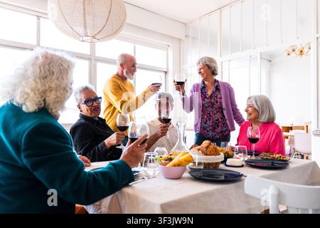 Gruppe lächelnder Seniorenfreunde, die eine fröhliche Dinnerparty zu Hause genießen, mit Rotwein anstoßen und Momente der Freundschaft und des Glücks teilen Stockfoto
