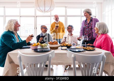 Sechs fröhliche Seniorenfreunde unterschiedlicher Ethnizitäten, die während eines geselligen Treffens zu Hause feiern und miteinander interagieren Stockfoto