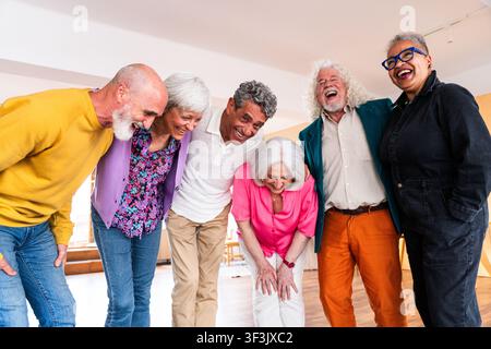 Verschiedene Gruppe von lächelnden Senioren, die einen freudigen Moment teilen, Freundschaft und Glück in einer entspannten Indoor-Umgebung umarmen Stockfoto