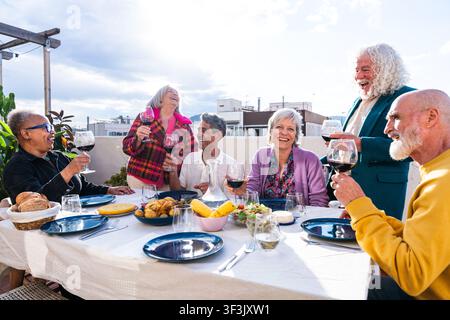 Gruppe lächelnder multikultureller Seniorenfreunde, die ein Mittagessen im Freien genießen und mit roten Weingläsern auf einem sonnigen Tag auf das Leben und die Freundschaft stoßen Stockfoto