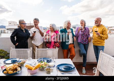 Gruppe vielfältiger Seniorenfreunde, die sich auf einer Dachterrasse zu einem fröhlichen Abendessen versammeln, lachen und Wein trinken, während sie ihre Freundschaft feiern und Stockfoto