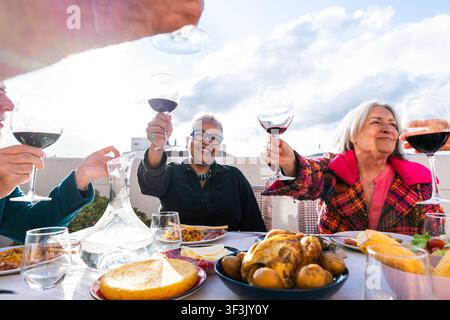 Gruppe verschiedener Senior-Freunde, die Weingläser heben, gemeinsam feiern und ein festliches Dachgeschichtessen mit verschiedenen Gerichten unter einem klaren Himmel genießen, Stockfoto