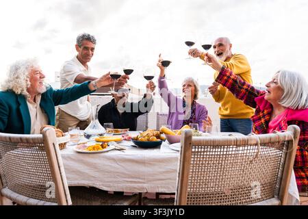 Verschiedene Gruppe von lächelnden Senioren, die sich auf einer Heimterrasse versammeln, Freundschaft und Lebensstil feiern und mit Rotwein anstoßen, während sie draußen sind Stockfoto
