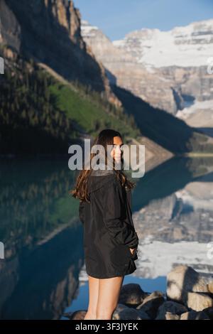 Ansicht einer jungen Frau, die am Ufer steht, mit schneebedeckten Bergen und ihrer Spiegelung im Wasser des Lake Louise in Alberta, Kanada. Stockfoto