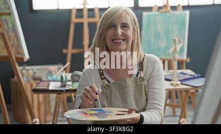 Frau Malerin im Atelier hält Palette und Pinsel, mischt mit der Hand helle Farben, lächelt im Schürzen und schaut in die Kamera, entspannte Haltung; kreativ Stockfoto