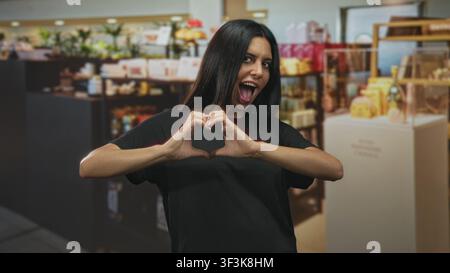 Frau, die mit Händen eine Herzform macht und lächelnd in Parfümvorstellungen in einem Ladengeschäft in Gebäuden steht; fröhliche Zuneigung. Stockfoto