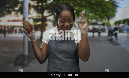 Frau hält Mango in behandschuhten Händen und verschränkt die Finger auf der Straße, trägt Denim-Schürze und weißes Polo; hoffe Frische. Stockfoto