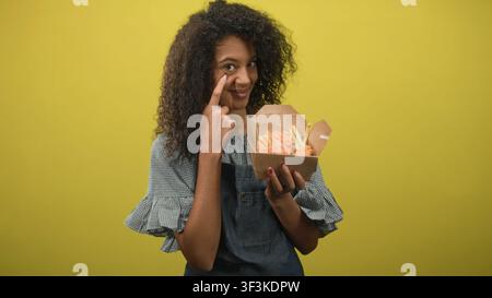 Teenager-Frau mit lockigem Haar in der Schürze zeigt Finger an Wange, während sie eine Kiste mit gebratenem Huhn und Pommes vor gelbem Studio-Hintergrund hält; S. Stockfoto