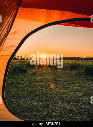 Orange Zelt öffnet sich und bietet einen atemberaubenden Blick auf die aufgehende Sonne über einem grünen Feld und einem fernen Wald Stockfoto