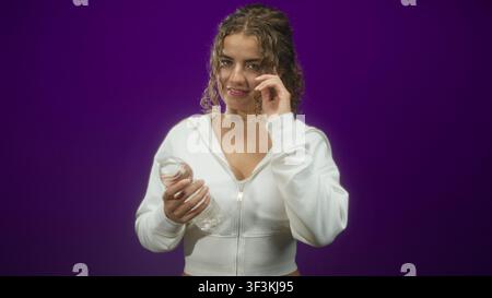 Frau, die eine Wasserflasche hält, die Hand an die Haare hält und mit dem Zeigefinger im Studio zeigt; Selbstvertrauen erfrischen. Stockfoto