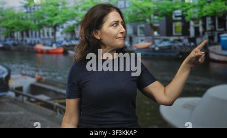 Die mittelalte hispanische Frau zeigt mit dem Finger nach rechts, die Augen sind fixiert, sie steht in der Nähe von vertäuten Booten und Ziegelgebäuden auf einer Straße mit marineblauen kurzen Ärmeln Stockfoto