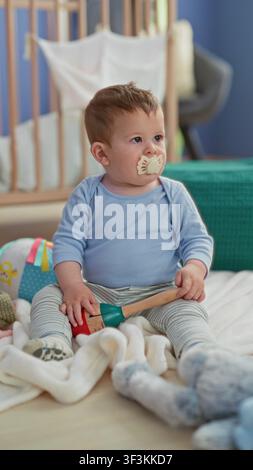 Kleinkind Junge, der auf einer Decke sitzt, Holzrassel hält und Plüschtier mit Schnuller im Mund in der Nähe eines Kinderbetts berührt; Neugier. Stockfoto