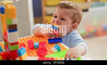 Junge kaut mit der Hand an einem bunten Plastikbaustein an einem Spieltisch und greift nach gestapelten Blöcken; Kindliche Neugier. Stockfoto
