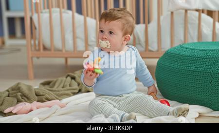Kleiner Junge hält ein farbenfrohes Ringspielzeug mit Schnuller im Mund, während er auf einer Decke in der Nähe des Kinderbetts im Kinderzimmer sitzt; Neugierde Komfort. Stockfoto