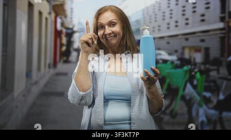 Ältere hispanische Frau, die eine blaue Seifenflasche hält, lächelt und zeigt den Zeigefinger nach oben auf eine Straße mit geparkten Fahrrädern und Gebäuden; Reinigung Stockfoto