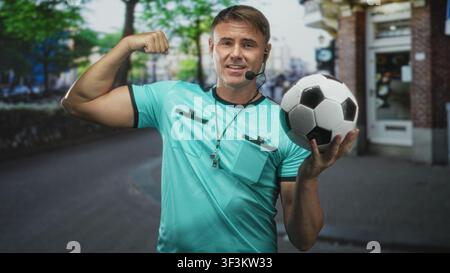 Mann Schiedsrichter im türkisfarbenen Hemd hält Fußball, trägt Pfeife und Headset, beugt den Arm und lächelt auf der Straße; Zuversicht. Stockfoto