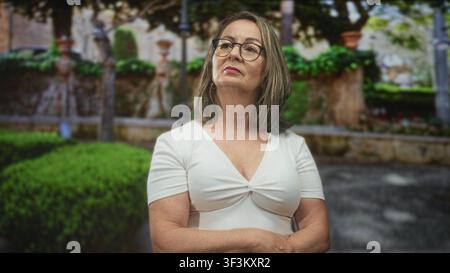 Hispanische Frau mit grauen Haaren, Brille, Arme überkreuzt und sichtbares Brustteil vor einem Steingebäude und Gartenschmuck; ruhige Ko Stockfoto