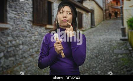 Junge chinesin, die Pinsel hält, Finger bis Kinn, Baskenmütze und lila Pullover auf der Kopfsteinpflasterstraße trägt; leises Denken. Stockfoto
