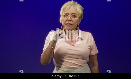 Eine ältere Frau zeigt mit dem Finger auf die Kamera, während sie im Studio rosa Polo, Brille, Armband und Ring trägt; ernste Warnung. Stockfoto