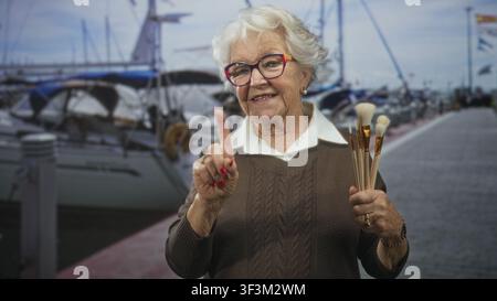 Eine ältere Frau mit Pinseln zeigt den Zeigefinger in Richtung des Betrachters, während sie auf einem Yachthafen neben verankerten Yachten steht; Selbstvertrauen. Stockfoto