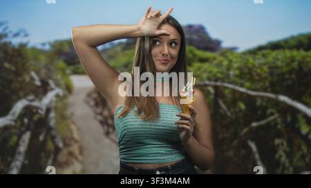 Die junge brünette Frau, die Eisbecher hält, lässt mich auf der Stirn ein Zeichen setzen, während sie auf einem Waldweg in der Nähe eines hölzernen Geländes steht; verspielter Sommerspaß. Stockfoto