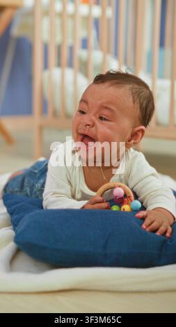 Hispanisches Baby, das eine Holzrassel hält, weint mit Tränen und Speichel, während der Betreuer sein Gesicht in einem Gebäude abwischt; Notfallbetreuung. Stockfoto
