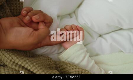 Kleiner Junge, dessen Hand den Finger eines Erwachsenen auf dem Bett umschließt, weißes Kissen und gestrickte Decke sichtbar; Bindungstendernheit. Stockfoto