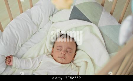 Baby schläft mit ausgestrecktem Arm und entspannter Hand in der Wiege im Kinderzimmer auf weißer Decke und geometrischem Kissen, Holzgeländer sichtbar; friedliche Ruhe. Stockfoto