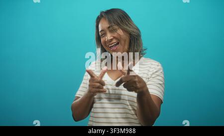 Frau zeigt beide Zeigefinger in Fingerpistolen, während sie im Studio mit türkisfarbener Kulisse lacht und lächelt; fröhliche Verspieltheit. Stockfoto