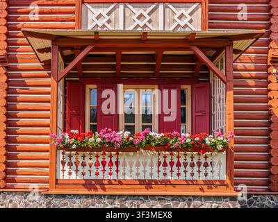 Eine bunte Blumenkiste auf einem kleinen Balkon umrahmt rote Holzläden eines traditionellen Blockhauses, das leuchtende Geranien, geschnitzte Geländerdetails und Stockfoto