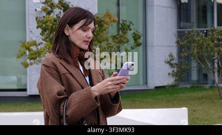 Eine reife Frau in ihren 50ern schaut nachdenklich in die Ferne, während sie ihr Handy hält. Betont Themen der Selbstreflexion, des Übergangs ins mittlere Lebensalter Stockfoto