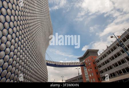 Breitwinklige, niedrigperspektivische Ansicht der ikonischen Selbstridges-Kaufhausfassade am Bullring, Birmingham, West Midlands, England, aufgenommen am 6. Mai 2012. Entworfen von Future Systems dominiert die futuristische Blobitecture-Struktur den linken Bildrahmen mit ihren silberfarbenen eloxierten Aluminiumscheiben, die über eine erhöhte Fußgängerbrücke mit dem mehrstöckigen Parkhaus Moor Street auf der rechten Seite unter einem hellen, teilweise bewölkten blauen Himmel verbunden sind. Stockfoto