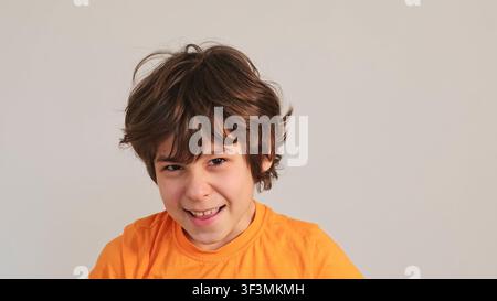 Nahaufnahme eines lächelnden Jungen in einem orangefarbenen Hemd, der gegen eine einfache Wand eine spielerische Grimasse schneidet, mit Platz für Text. Kindheitsgefühle, Selbstvertrauen und Stockfoto