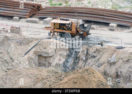 Betonpumpmaschine auf Schotterboden, tragbare Pumpe mit verrosteter, verwitterter Optik, Schlauch führt zum Betonbereich, Haufen aus Sand, Kies und Betonstahl Stockfoto