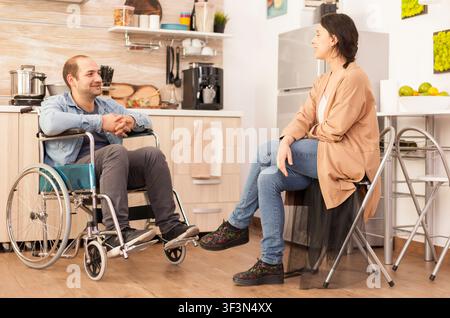 behinderter Ehemann und Ehefrau lächeln sich in der Küche an und zeigen Beziehungsunterstützung, Mobilitätsbehinderung und tägliche Heimintegration. Stockfoto
