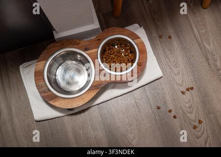 Edelstahl-Haustierschüsseln in einer Holzhalterung auf einem hellen Boden, mit Wasser, Trockenfutter und verstreutem Trockenfutter, die einen gemütlichen Fütterungsbereich schaffen Stockfoto