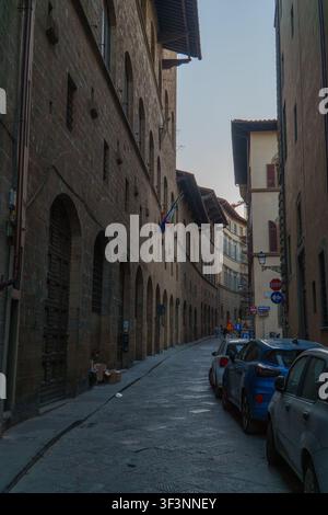 Florenz, Toskana, Italien. 28. Mai 2026. - Eine schmale, sonnenbeschienene Kopfsteinpflastergasse im historischen Florenz, Italien, gesäumt von alten Steingebäuden, geparkte V Stockfoto