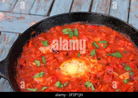 Traditionelle Shakshuka mit pochierten Eiern in würziger Tomatensoße, serviert in einer gusseisernen Pfanne auf einem blauen Holztisch Stockfoto