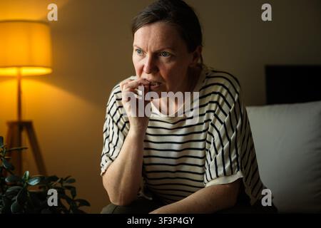 Frau, die sich die Nägel beißt, während sie zu Hause mit Stress und ängstlichen Gedanken kämpft. Bewusstsein für psychische Gesundheit, emotionales Wohlbefinden und Alltagsleben konzipiert Stockfoto