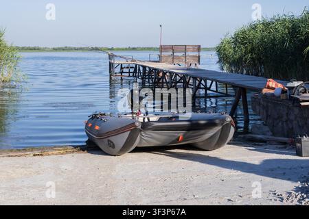 Aufblasbares Fischerboot mit Außenbordmotor, das neben einem Holzsteg an einem ruhigen Süßwassersee für die Freizeitfischerei bereit liegt. Stockfoto