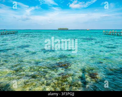 Klares türkisfarbenes Meerwasser, das über Korallenriffe und weißen Sand fließt, mit zwei hölzernen Anlegestegen, die sich unter einem hellen Himmel in Karimun ins Meer erstrecken Stockfoto