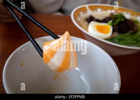 Chopsticks heben einen weichen, orange-weißen Cedea-Käse-Dumpling-Fischkuchen an. Die Schüssel enthält halb gegessenes Ei und Grünzeug. Das Essen steht auf einem warmen Holztisch. Moment Stockfoto