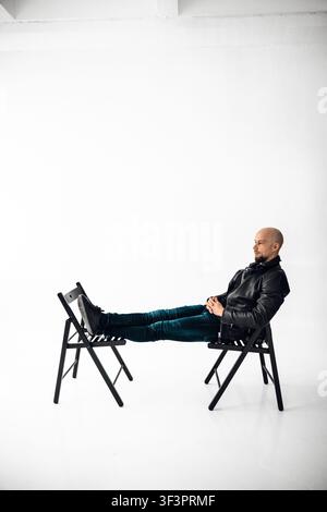 Mann sitzt auf einem Stuhl mit ausgestreckten Beinen auf einem anderen Stuhl in einem einfachen Studio Stockfoto