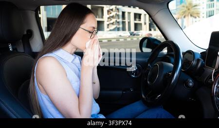 Junge Frau, die ihre Nase mit einem Taschentuch bedeckt, während sie in einem Auto sitzt. Konzept für saisonale Allergien, Erkältungssymptome und Atemwegspflege. Stockfoto
