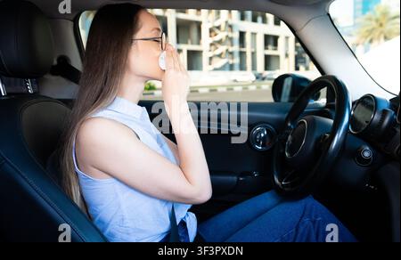 Junge Frau sitzt in einem Auto und niest in ein Taschentuch. Konzept für saisonale Allergie, Erkältungssymptome und Atemwegengesundheit mit natürlicher Lebensstilumgebung. Stockfoto